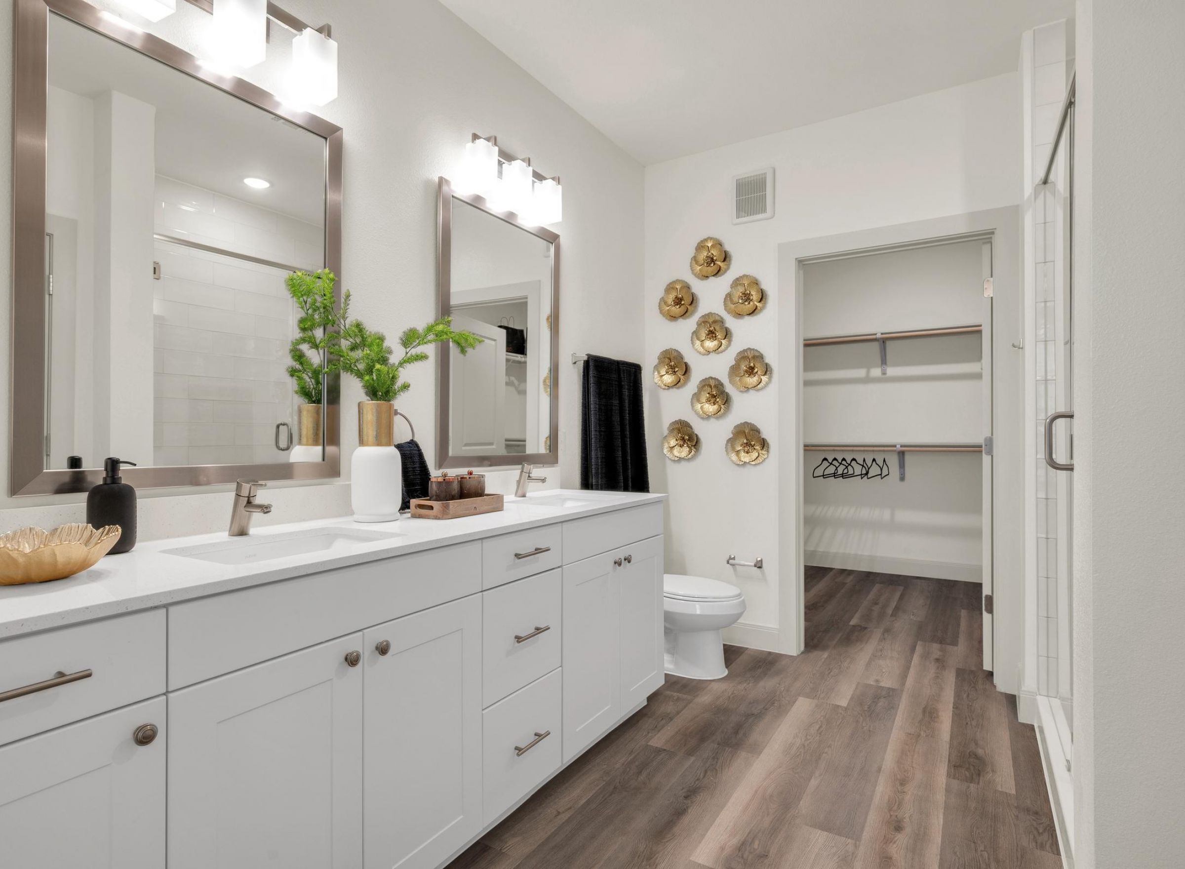 Camber Ridge Modern bathroom with dual sinks, wall decor, and open closet. White cabinets, wooden floor, and bright lighting.