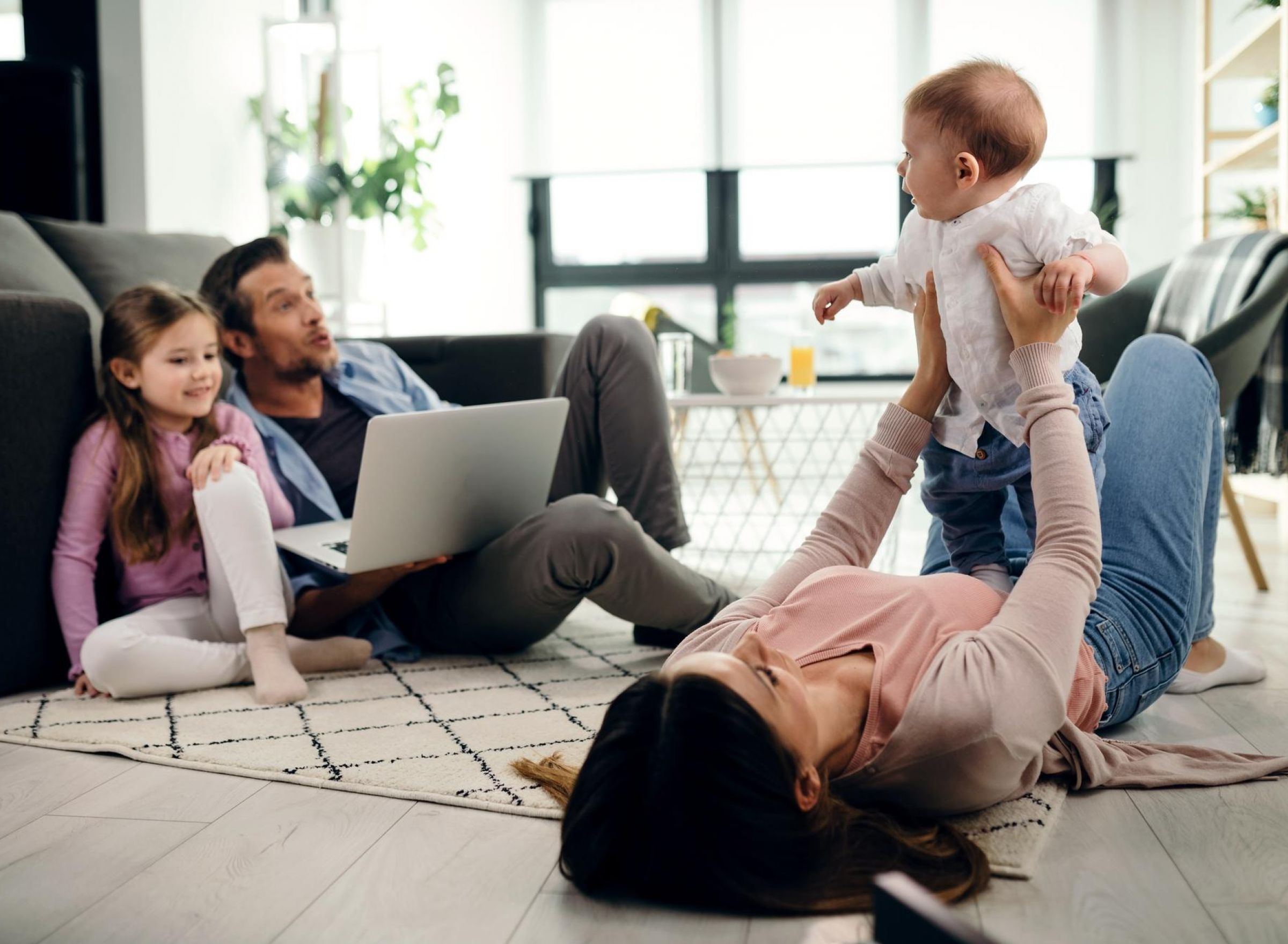 Camber Ridge Family enjoying time together at home: a woman plays with a baby, while a man and girl sit nearby with a laptop.