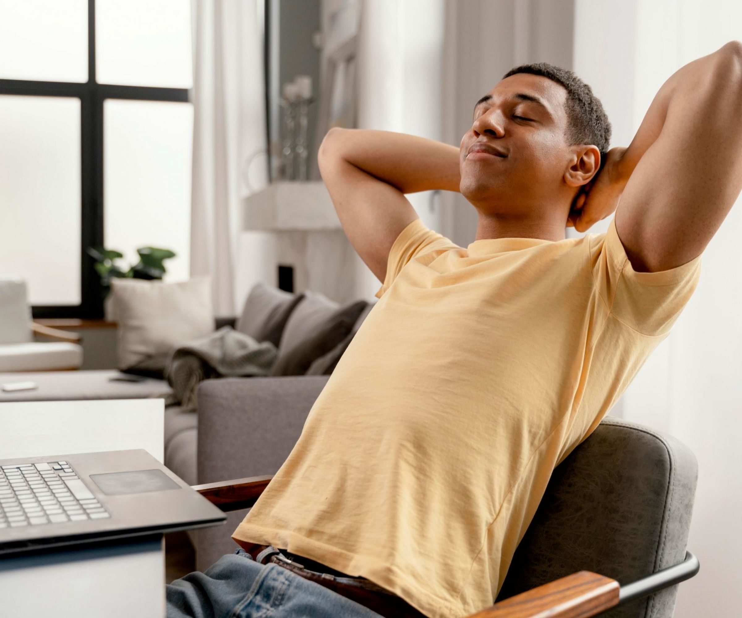 Camber Ridge Man in yellow shirt relaxing with hands behind head at desk, laptop open in bright, modern room.