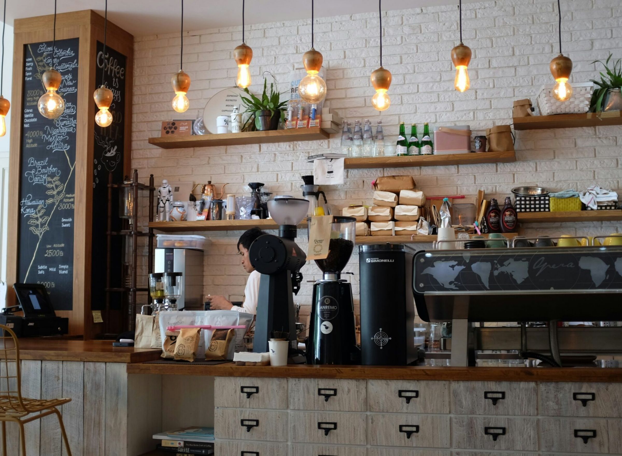 Camber Ridge Trendy coffee shop interior with hanging lights, plants, a chalkboard menu, and espresso machines on a wooden counter.